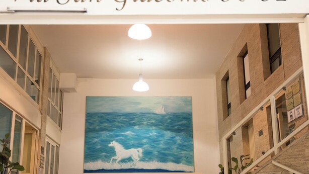 Un couloir orné d’un tableau représentant un cheval et des vagues, une enseigne indiquant « Via San Giacomo 30-32 », et un escalier sur la droite.