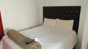 A neatly made bed with a rolled towel and remote control, a plain white wall, and a simple headboard with buttons.