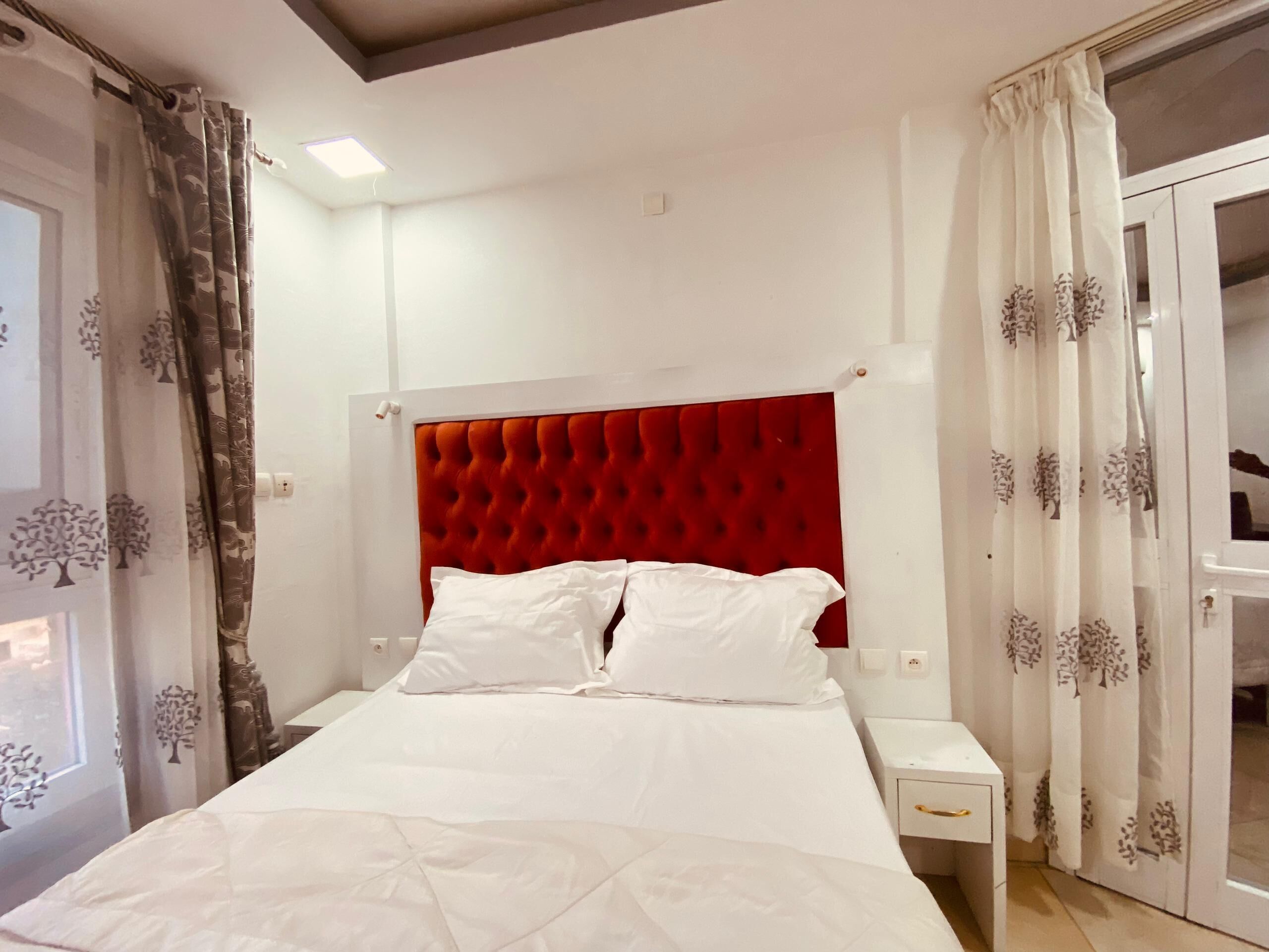Habitación de hotel con cabecera tapizada en rojo, ropa de cama blanca y puerta con vistas.