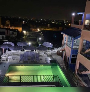 Un área de piscina en la azotea con múltiples sillones de descanso y sombrillas, rodeada de edificios y luces de la ciudad al fondo.