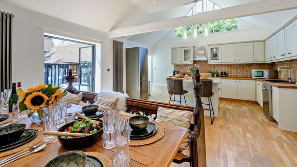 A spacious kitchen with white cabinets, a dining area with a wooden table set for a meal, and a view of the outside through large windows. - The Willows -  a holiday let by Bloom Stays that sleeps 8 guests  in 4 bedrooms (Ash)