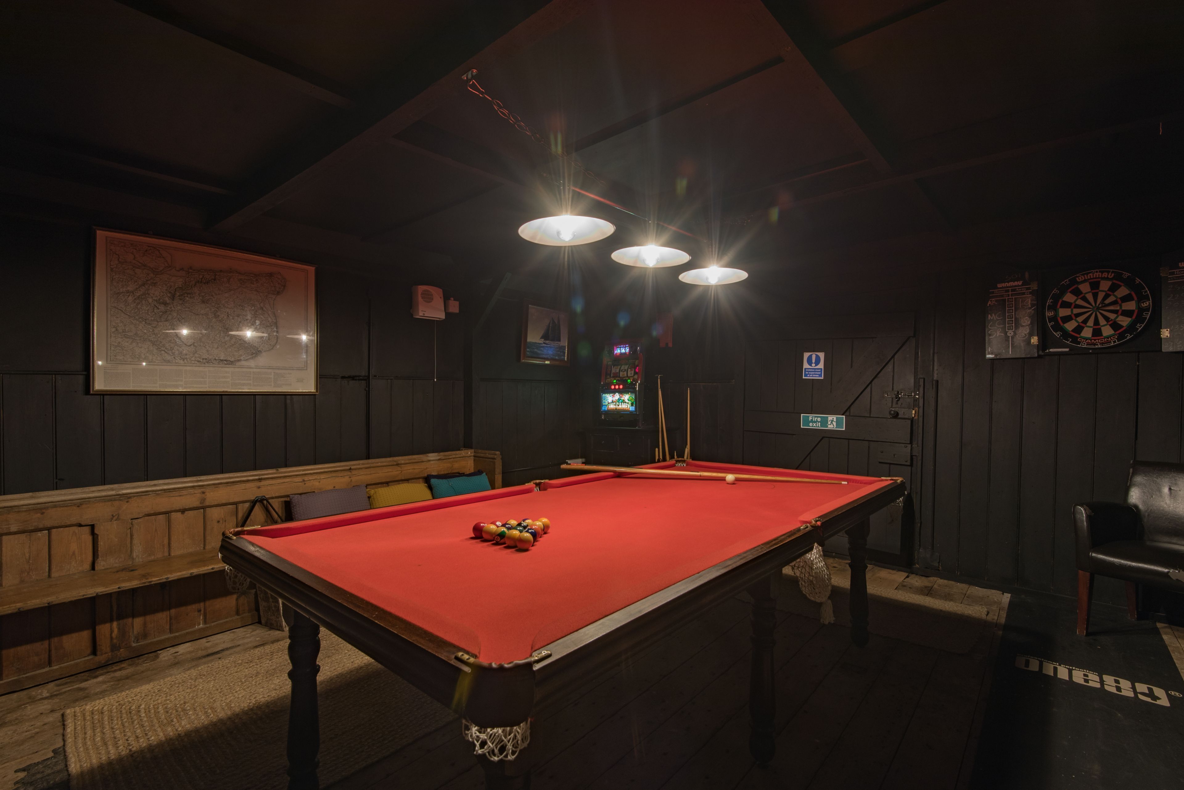 A billiards table with a red cloth, a wooden bench, and a dartboard on the wall.