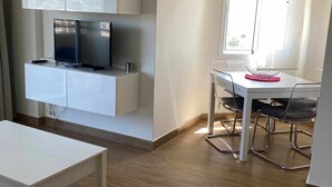 Een moderne, goed verlichte kamer met een flatscreen-televisie bevestigd aan een wit kastje, een eettafel met stoelen en een raam met zonwering.