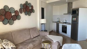Een moderne woonkamer met een beige bank, een decoratief plaid en een klein, rond tafeltje. Op de achtergrond is een keukenhoek zichtbaar met een magnetron, oven en spoelbak.