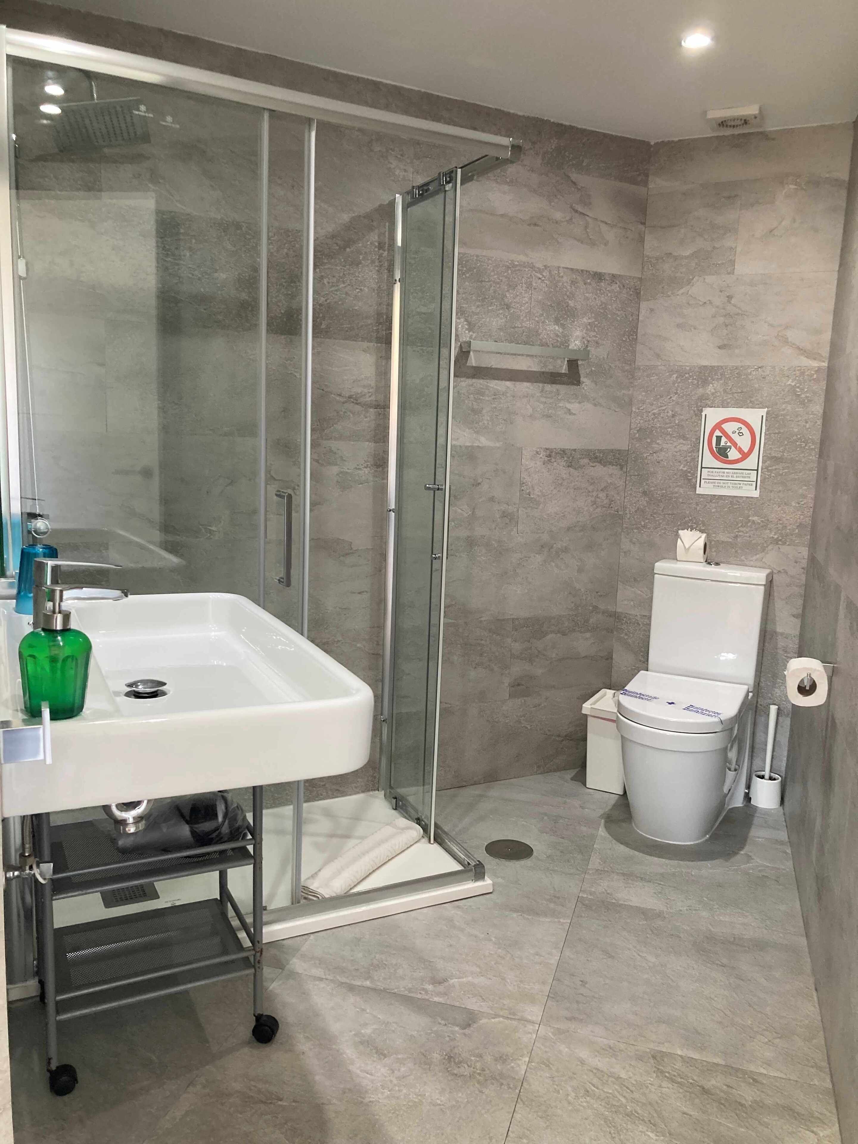 A modern bathroom with a glass-enclosed shower, a white sink with a green soap dispenser, and a toilet with a paper towel holder.