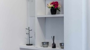 A white interior wall with three shelves. The top shelf holds a small potted plant, a decorative bowl, and a small pot. The middle shelf has a vase with pink and white flowers. The bottom shelf features a black candle holder, a small bowl, and a bird figurine. - [Lingotto - Inalpi Arena] Bright Apartment (Torino)