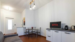 A modern living room with a sofa, a dining area with a table and chairs, a kitchen with a TV displaying 'NETFLIX', and a coffee machine. - [Lingotto - Inalpi Arena] Bright Apartment (Torino)
