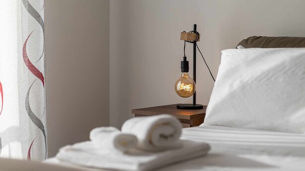 A neatly made bed with white linens, a wooden nightstand with a vintage-style lamp, and a rolled towel on the bed. - [Lingotto - Inalpi Arena] Bright Apartment (Torino)