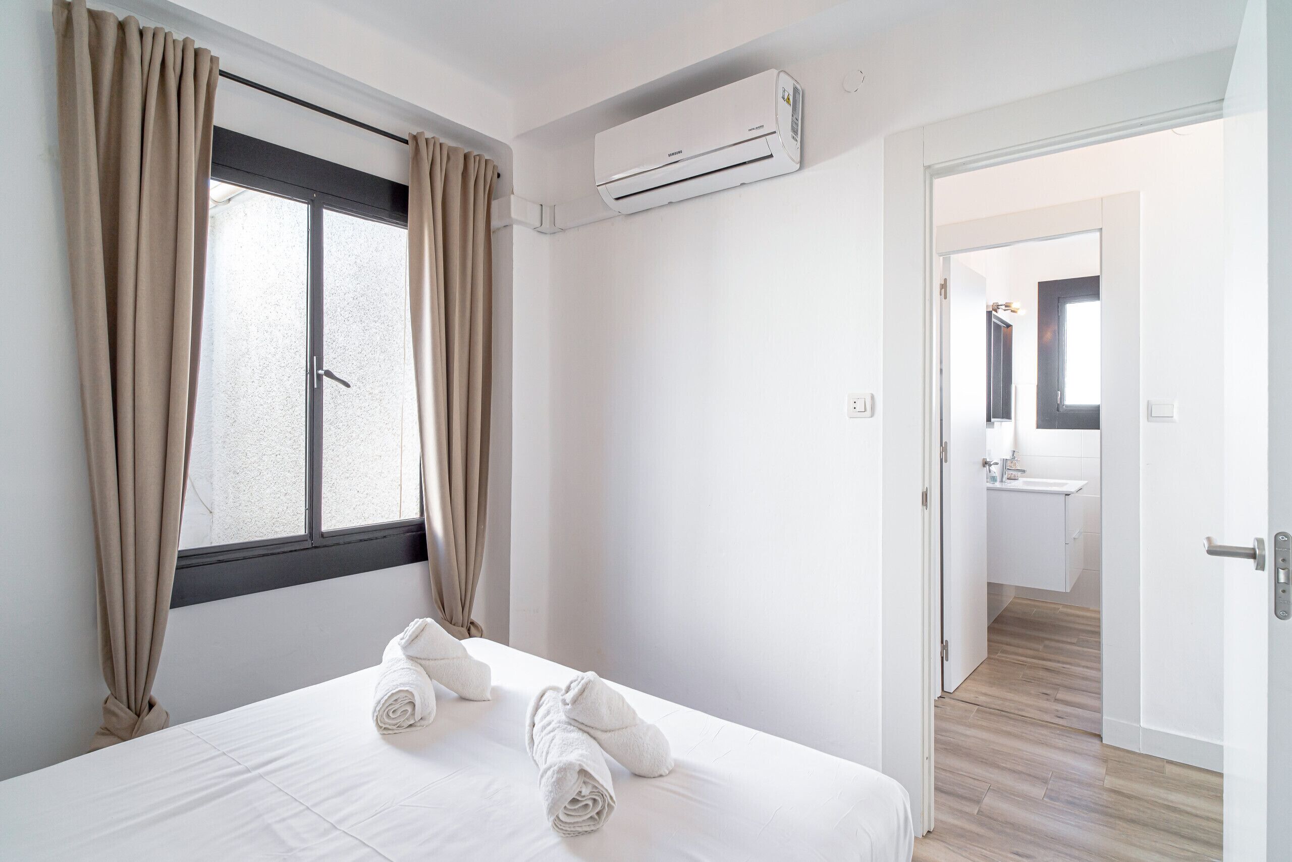 A hotel room with a bed, beige curtains, an air conditioning unit, and a view of a bathroom.