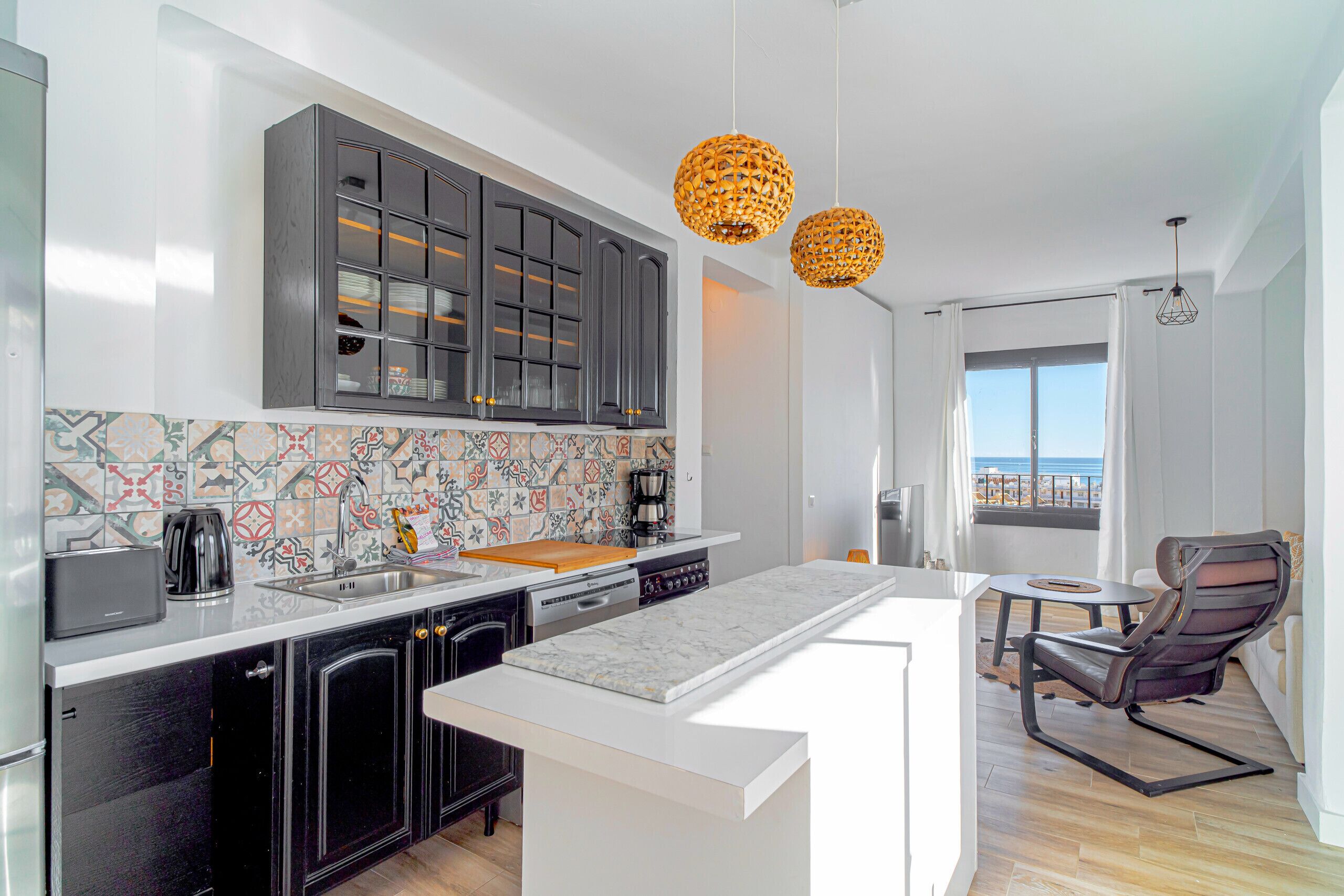 Una cocina moderna con una isla grande, armarios oscuros y un salpicadero colorido. Hay un comedor con una mesa y sillas, y una vista al mar a través de una ventana.