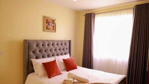 A hotel room with a tufted headboard bed, white linens, red and white pillows, a wooden side table, and a framed picture on the wall. - Lux Suites Greatwall Getaway Apartments (Athi River)