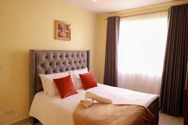 Een hotelkamer met een bed met gestuikt hoofdbord, witte lakens, rode en witte kussens, een houten nachtkastje en een lijst met een afbeelding aan de muur.