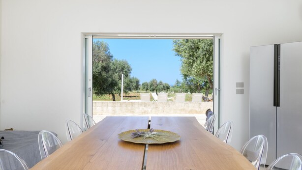 A modern dining area with a wooden table, clear chairs, and a view of a garden through a glass door. - Villa La Guaceta by Wonderful Italy (Carovigno)