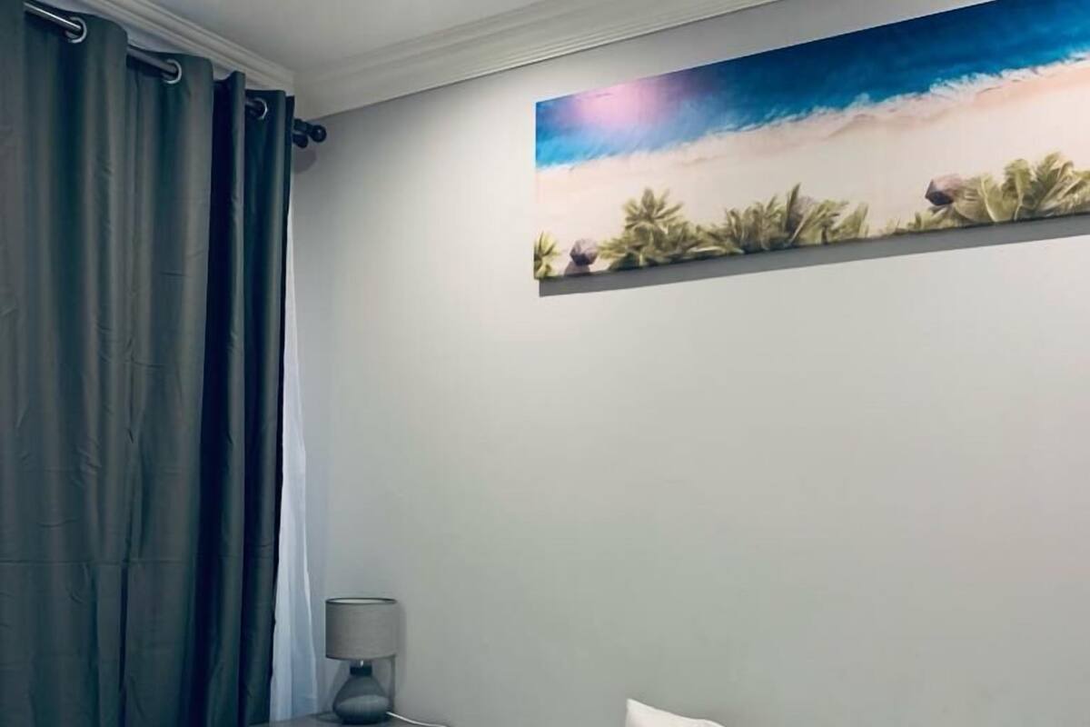 Una habitación de hotel con una cama, una mesita de noche con una lámpara y un cuadro de un paisaje tropical en la pared.