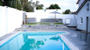 A rectangular swimming pool with a clear blue water surface, surrounded by a white wall and a grey deck area with a small grassy patch and a bench. - Semeni Asante Getaway (Johannesburg South)