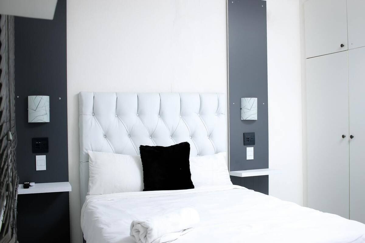 Una cama bien hecha con sábanas blancas y un almohadón negro, un cabecero blanco acolchado y un armario gris a la derecha.