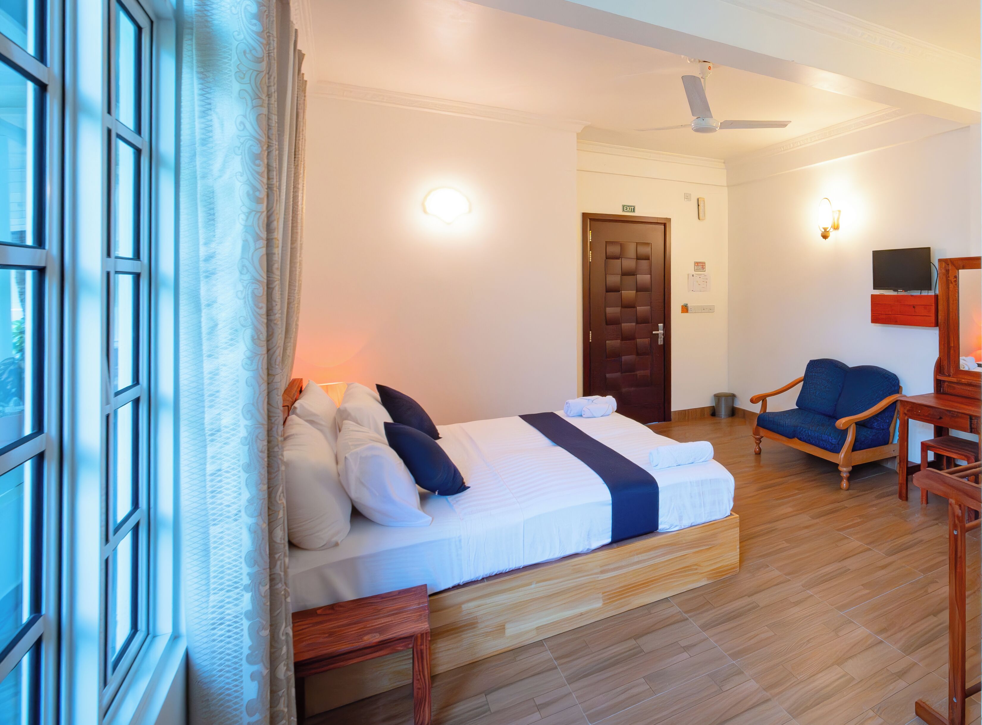 Une chambre d’hôtel avec un lit, une chaise, un plancher en bois, un ventilateur de plafond et une porte avec un panneau indiquant la sortie.