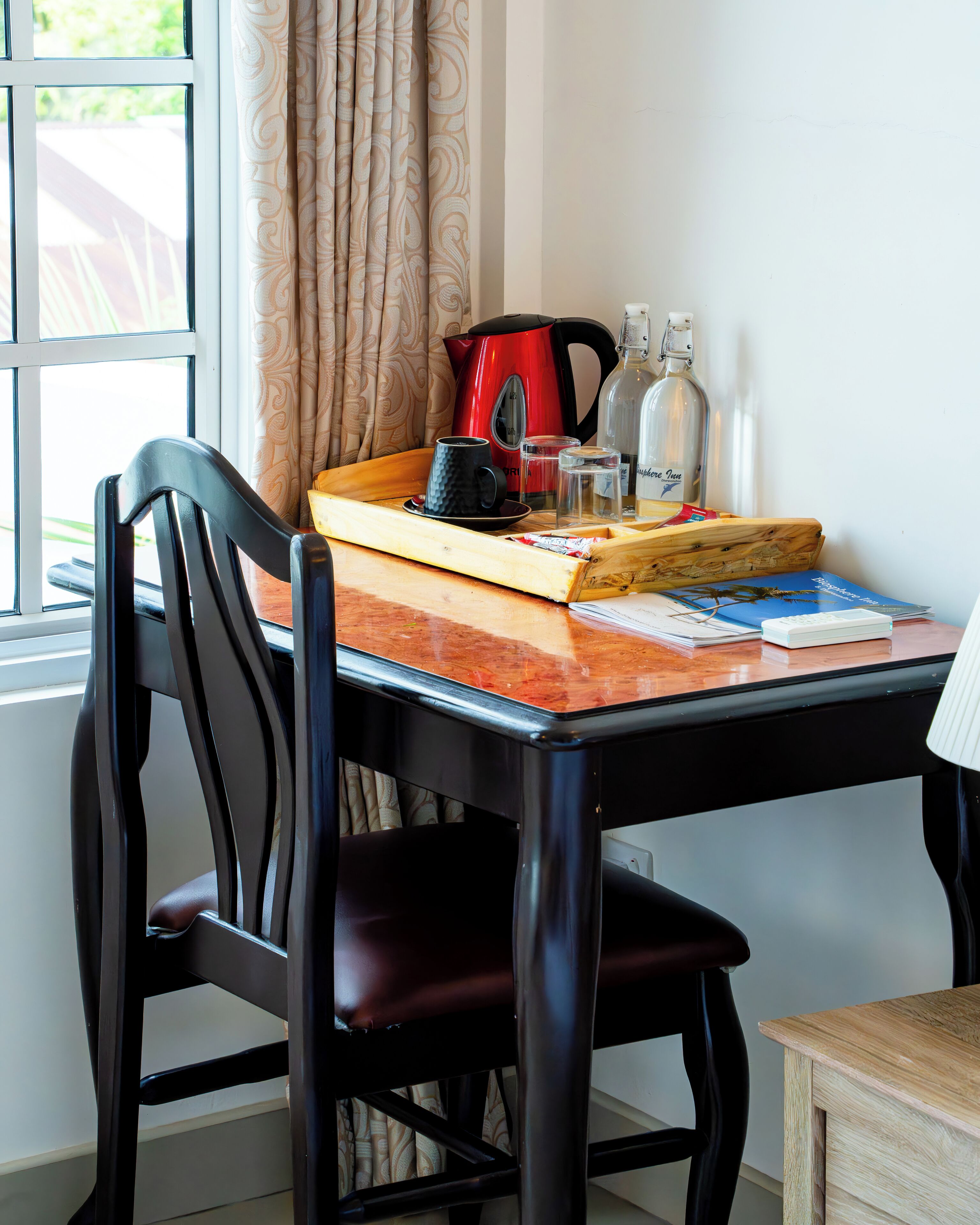 Un coin repas avec une table en bois, des chaises noires, un plateau sur lequel se trouvent une tasse noire, une bouilloire rouge et une bouteille d’eau.