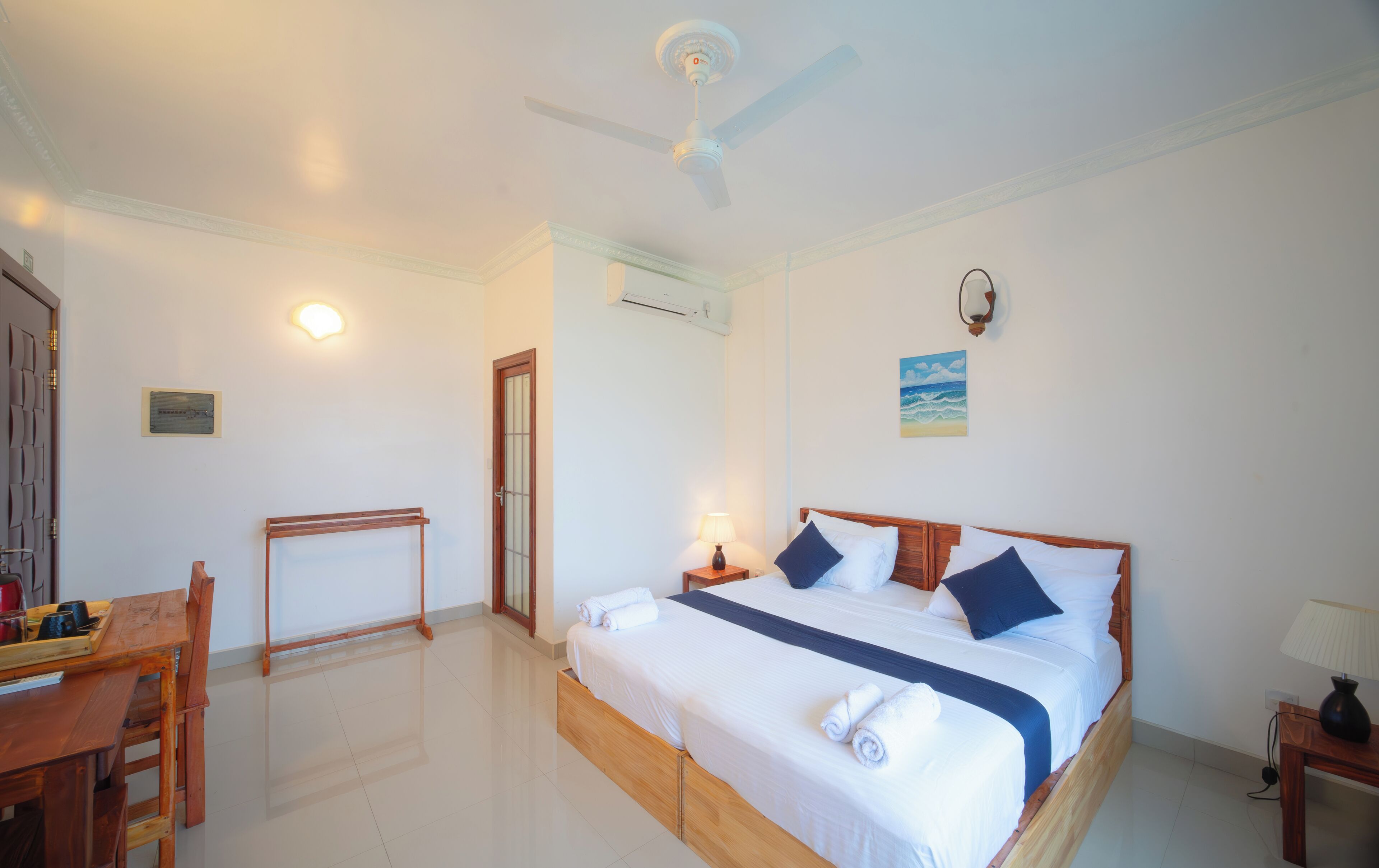 A hotel room with a bed, a desk, a chair, a ceiling fan, and a painting on the wall.