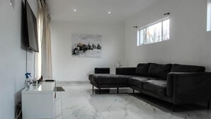 Un salon moderne avec un canapé d’angle, un téléviseur à écran plat fixé au mur, une fenêtre avec des stores et un sol en marbre.