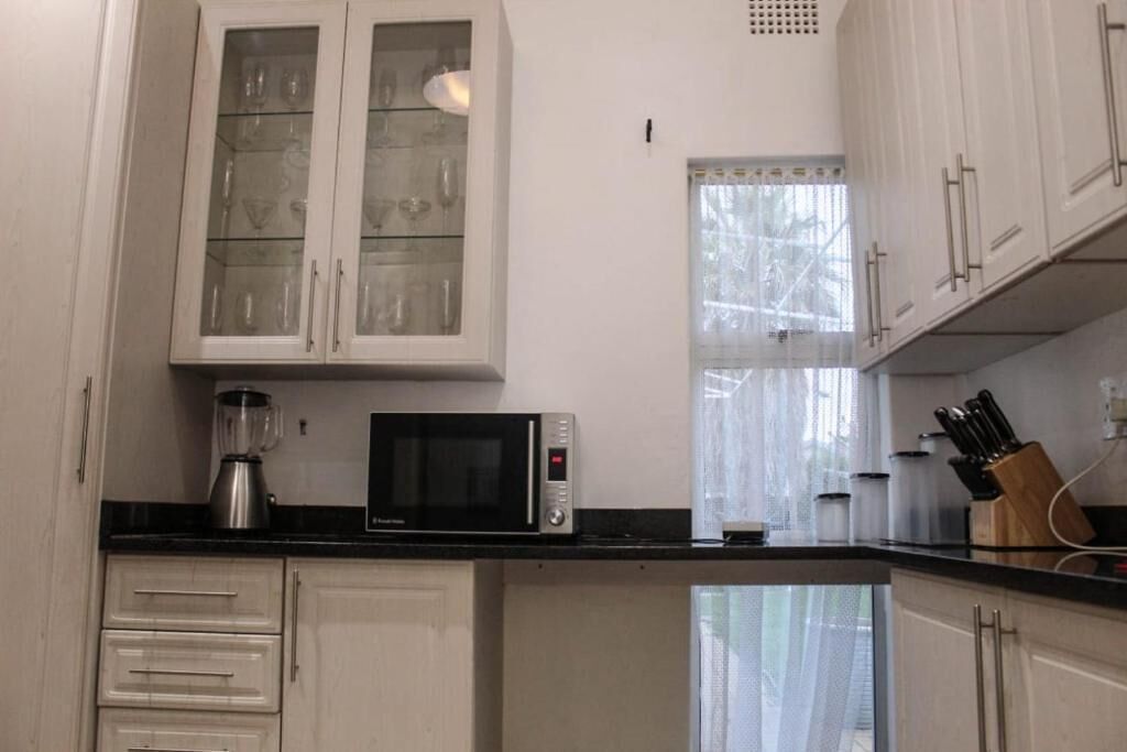 A kitchen with white cabinets, a microwave, a blender, and a window with sheer curtains.