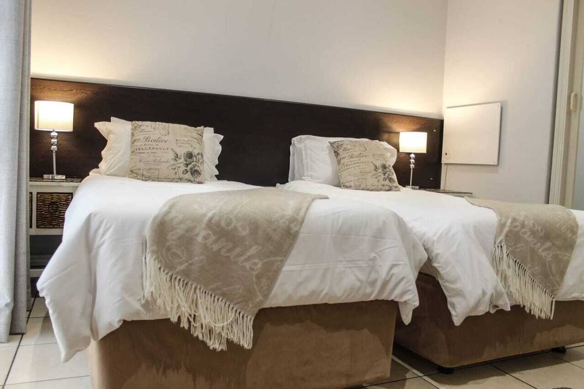 A double bed with white linens and two decorative pillows, a bedside table with a lamp, and a wall-mounted light fixture.