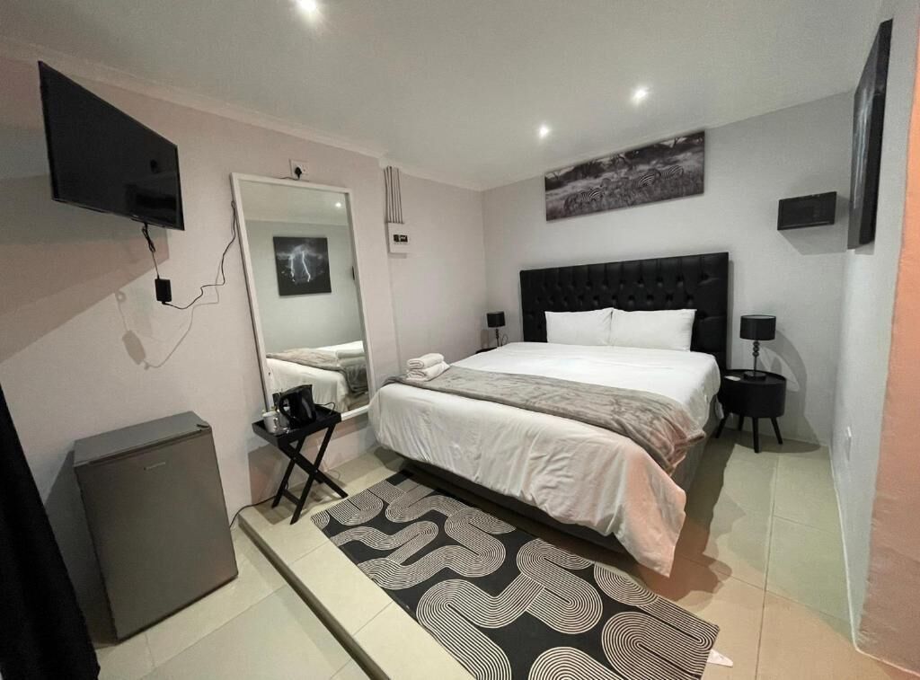 A modern bedroom with a large bed, a flat-screen TV mounted on the wall, a small refrigerator, and a mirror.