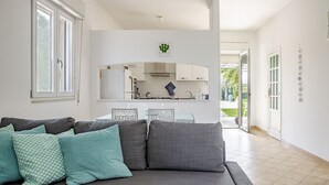 A modern living room with a grey sofa, teal patterned cushions, a kitchen in the background, and a view of the outdoors. - Villa Bouganville by Wonderful Italy (Syracuse)