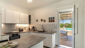 A modern kitchen with white cabinets, a countertop, and a view of an outdoor pool area. - Villa Bouganville by Wonderful Italy (Syracuse)