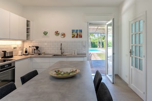 Une cuisine moderne avec des meubles blancs, une table Ă manger avec des chaises et une vue sur une piscine par une porte ouverte.