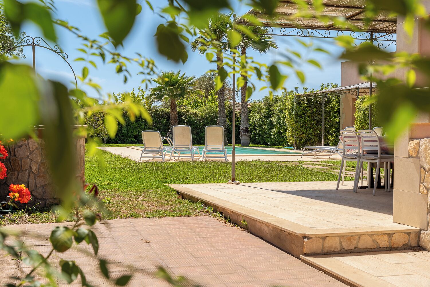 A poolside area with lounge chairs and a table, surrounded by greenery and a clear blue sky.