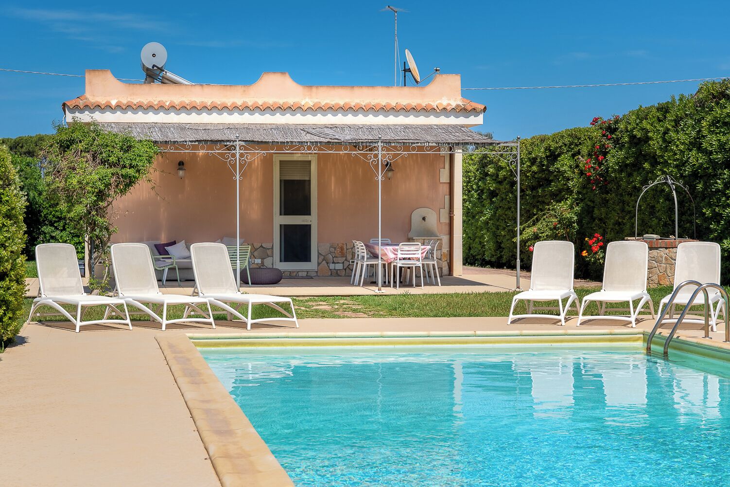 A pool area with lounge chairs and a table, a building with a tiled roof, and satellite dishes.