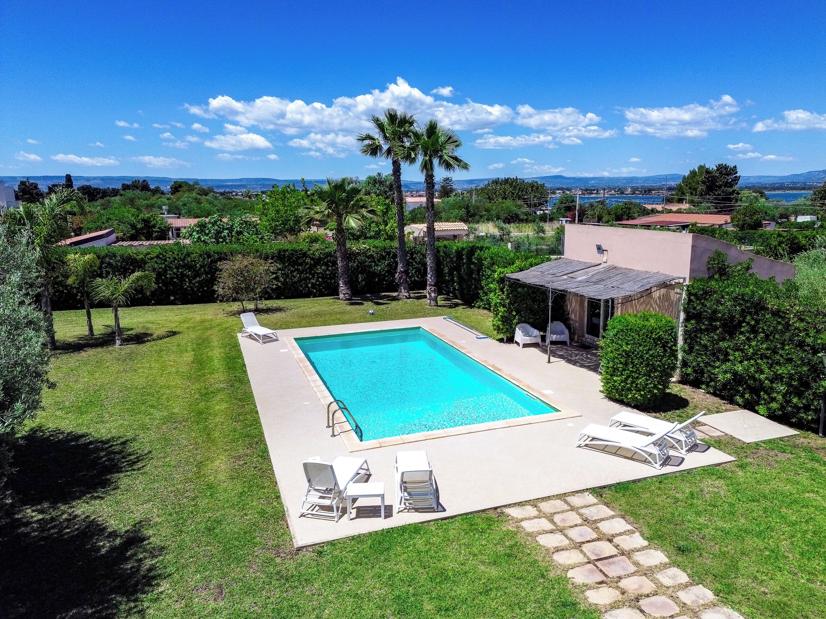 A rectangular swimming pool with a clear blue water, surrounded by a paved deck, and equipped with several lounge chairs.