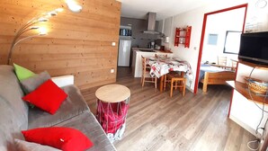 Un ambiente compatto con angolo cottura, zona pranzo, divano con cuscini, poggiapiedi a tamburo e televisione.