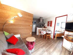 A compact living space with a kitchenette, a dining area, a sofa with cushions, a drum stool, and a television.