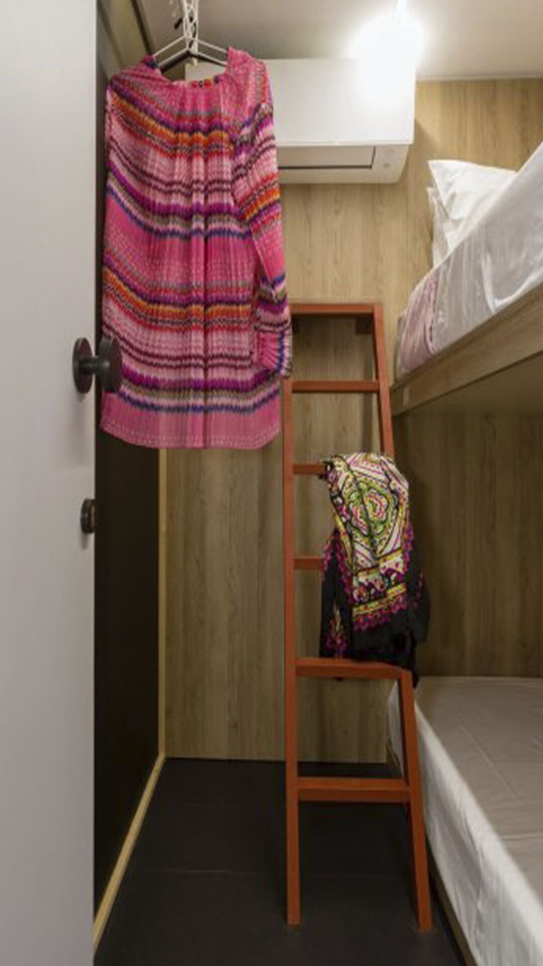 A small, compact room with a bunk bed, a wooden ladder, and a hanging rack with colorful clothing.