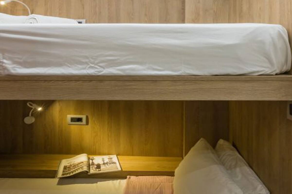A bunk bed with a white mattress and a lower bed with a pink blanket. There is a book on the lower bed and a wall-mounted light fixture above it.