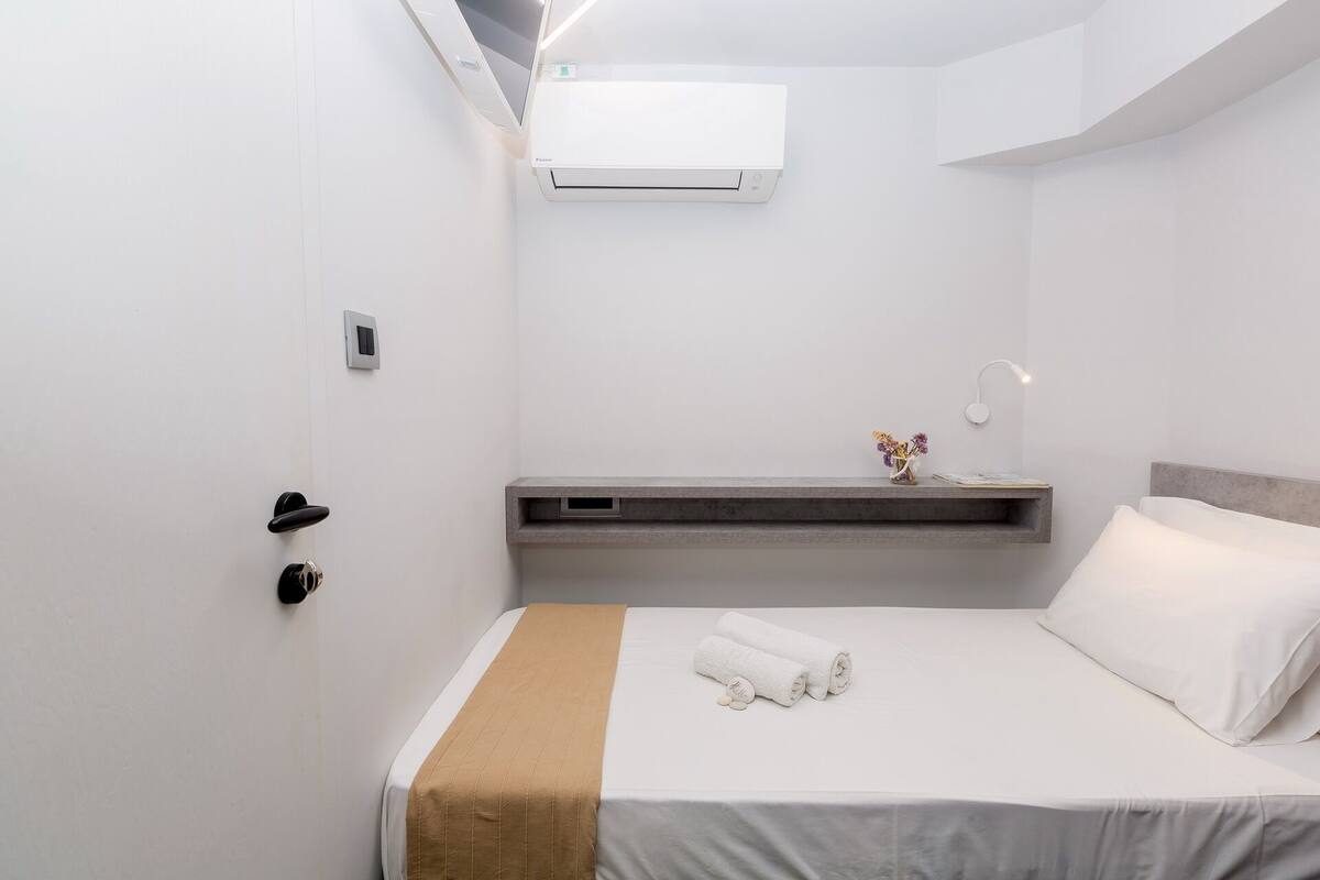A hotel room with a bed, a bedside table with a lamp, a small shelf, and an air conditioning unit on the wall.