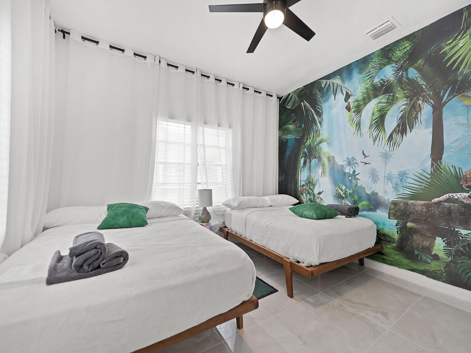 A bedroom with two beds, white bedding, green pillows, a ceiling fan, and a mural of a jungle scene.