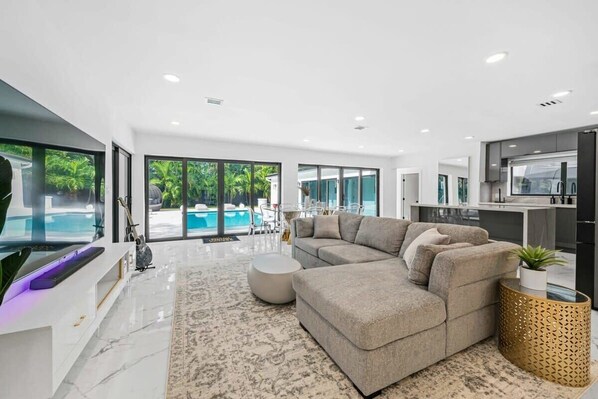 A modern living room with a sectional sofa, a flat-screen TV, and a large window overlooking a pool. - Luxury Villa Azure with Heated Pool 20min to Beach (Miami)