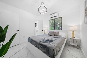A modern bedroom with a large bed, white walls, a ceiling light, and a window with blinds.