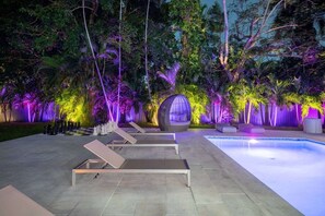 A pool area with lounge chairs and a small seating area surrounded by lush greenery and purple lighting.