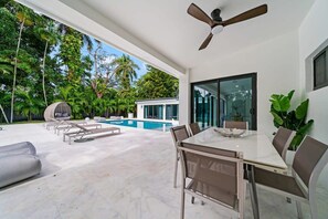 A modern outdoor patio with a glass table and chairs, a ceiling fan, and a pool.