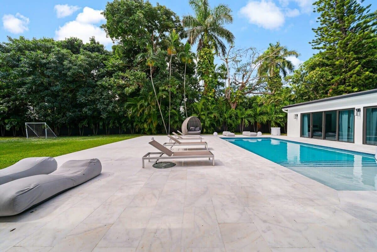 A modern outdoor pool area with lounge chairs and a clear blue pool, surrounded by lush greenery and palm trees.