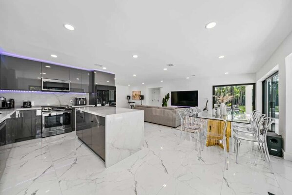 A modern kitchen with a central island, a dining area with clear chairs, and a large TV. - Luxury Villa Azure with Heated Pool 20min to Beach (Miami)