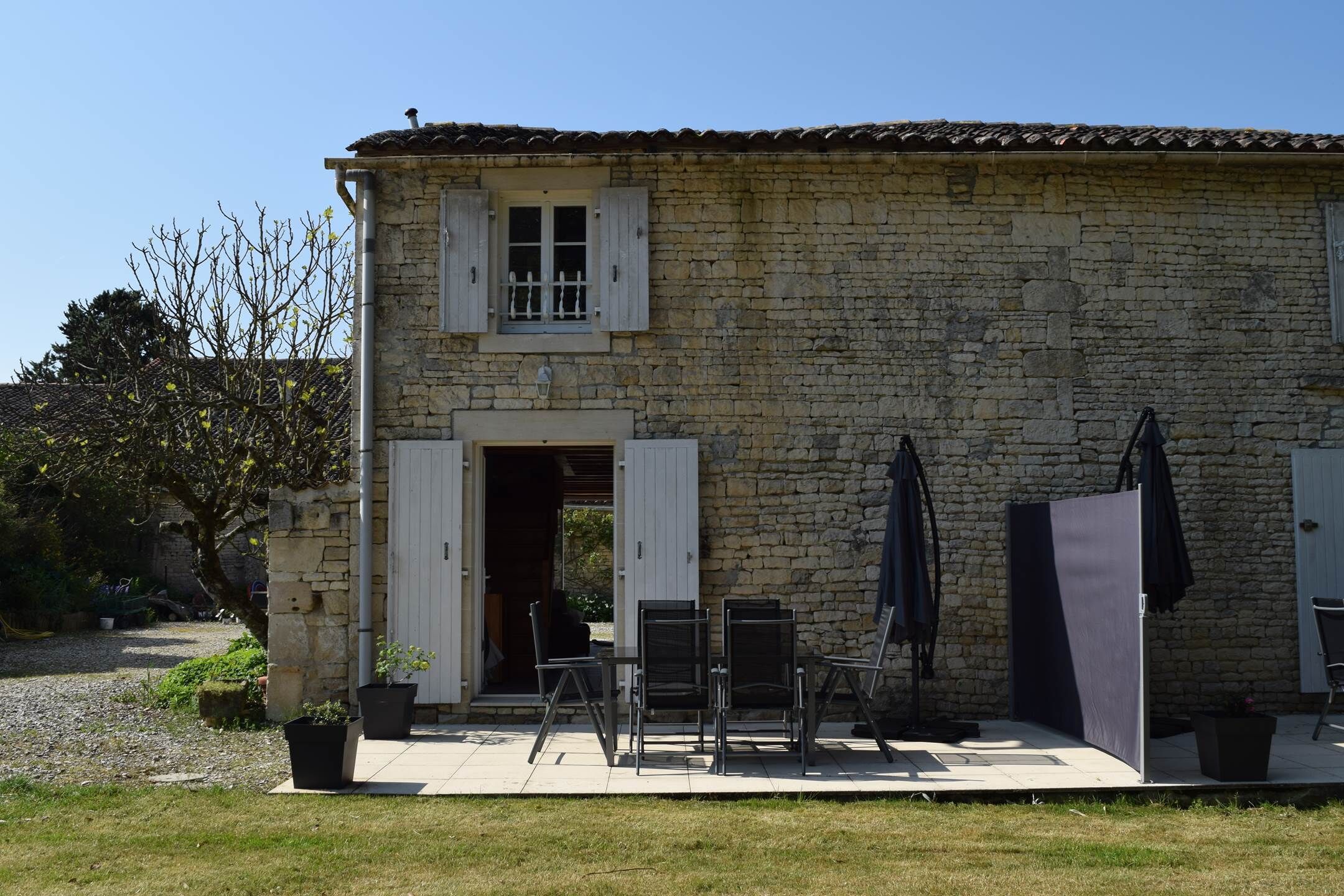 Ein Steinhaus mit einer Terrassenfläche, ausgestattet mit einem Tisch und Stühlen, einer Tür und einem Fenster mit Rollläden.