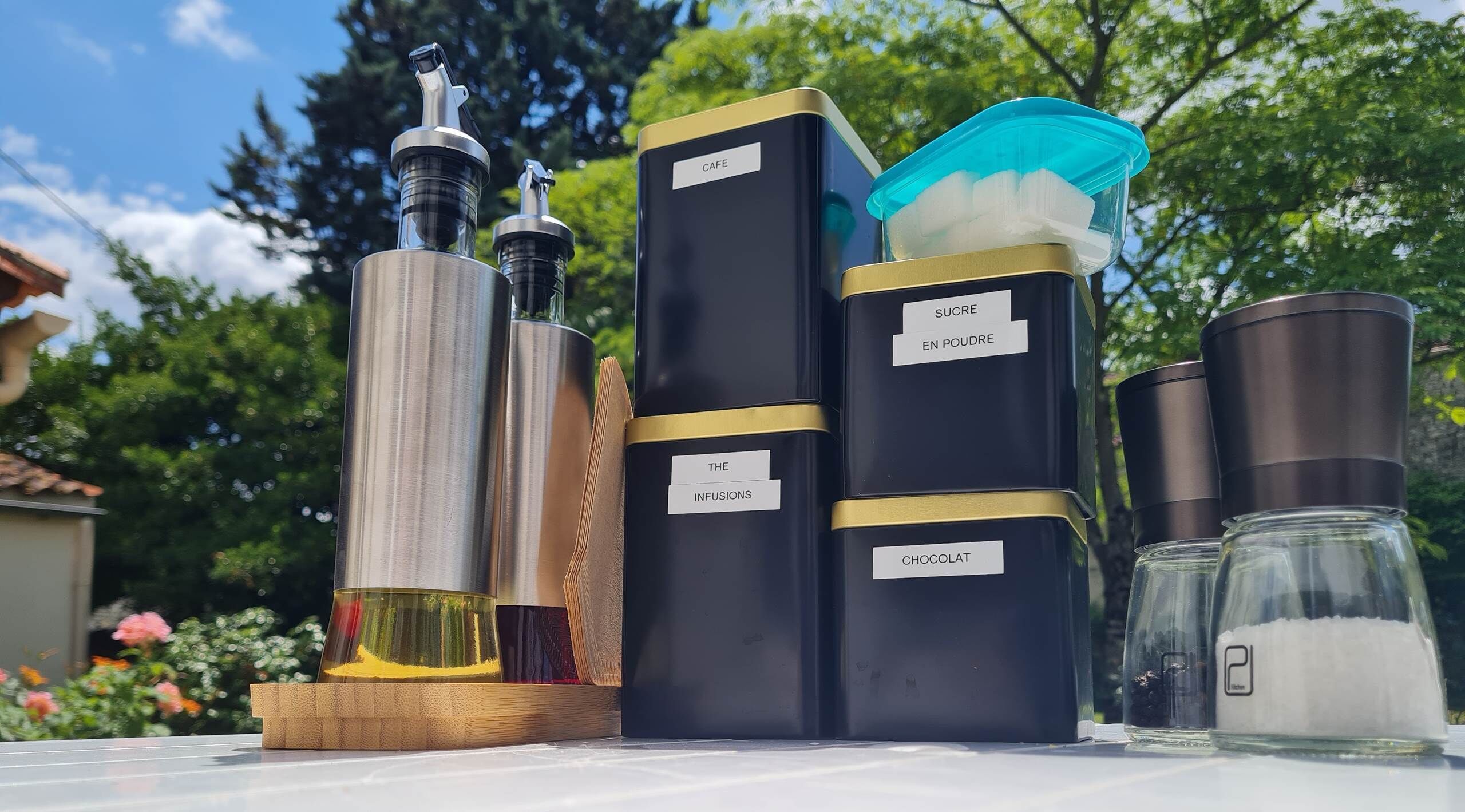 A collection of containers with labels on a table outdoors, surrounded by greenery.