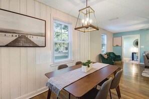 A dining area with a wooden table, chairs, a hanging light fixture, and a large framed picture on the wall.