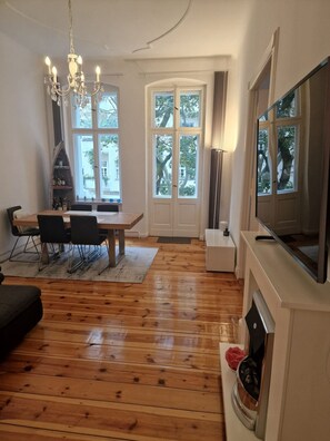 Een woonkamer met houten vloer, een eettafel met stoelen, een kroonluchter, een televisie en een open haard.