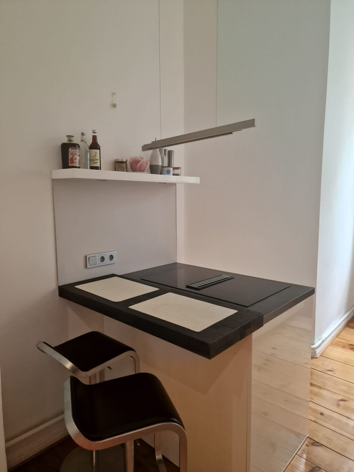 A compact kitchenette with a black countertop, two white placemats, a stainless steel chair, and a white shelf with bottles and a vase.
